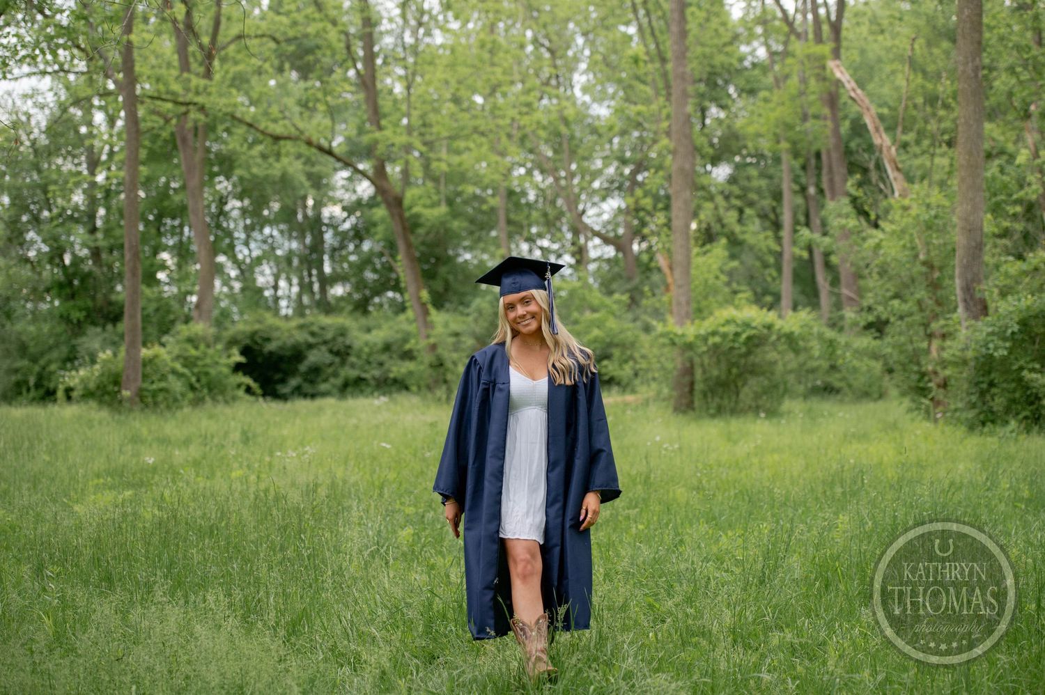 Graduate stands in a green spring meadow wearing traditional cap and gown against a backdrop of lush trees.