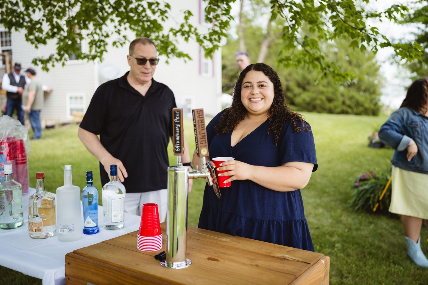 Two people serve drinks at an outdoor bar setup with various bottles on a wooden table during a summer event.