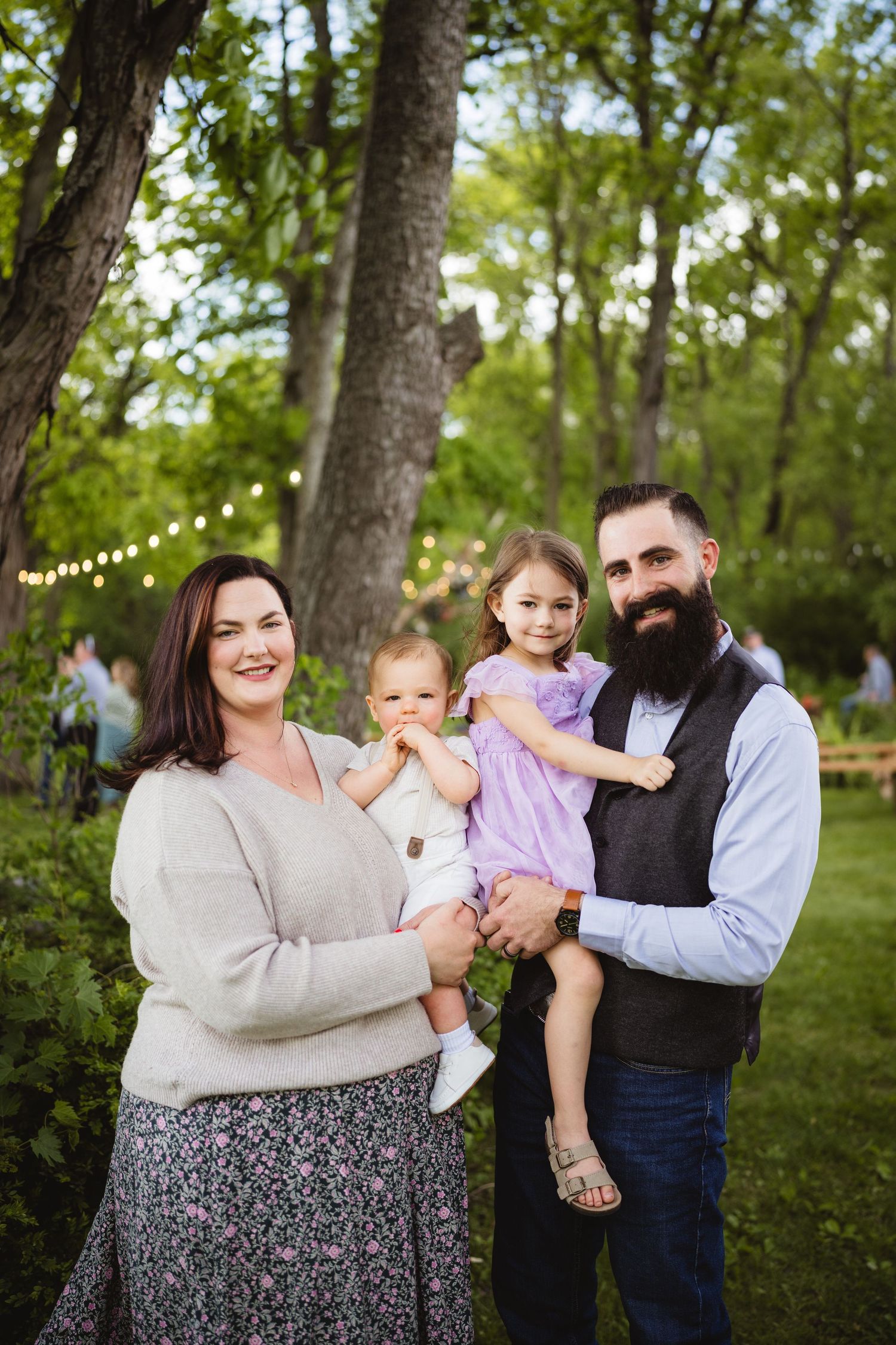 A family stands together outdoors in a garden setting with string lights visible in the background.