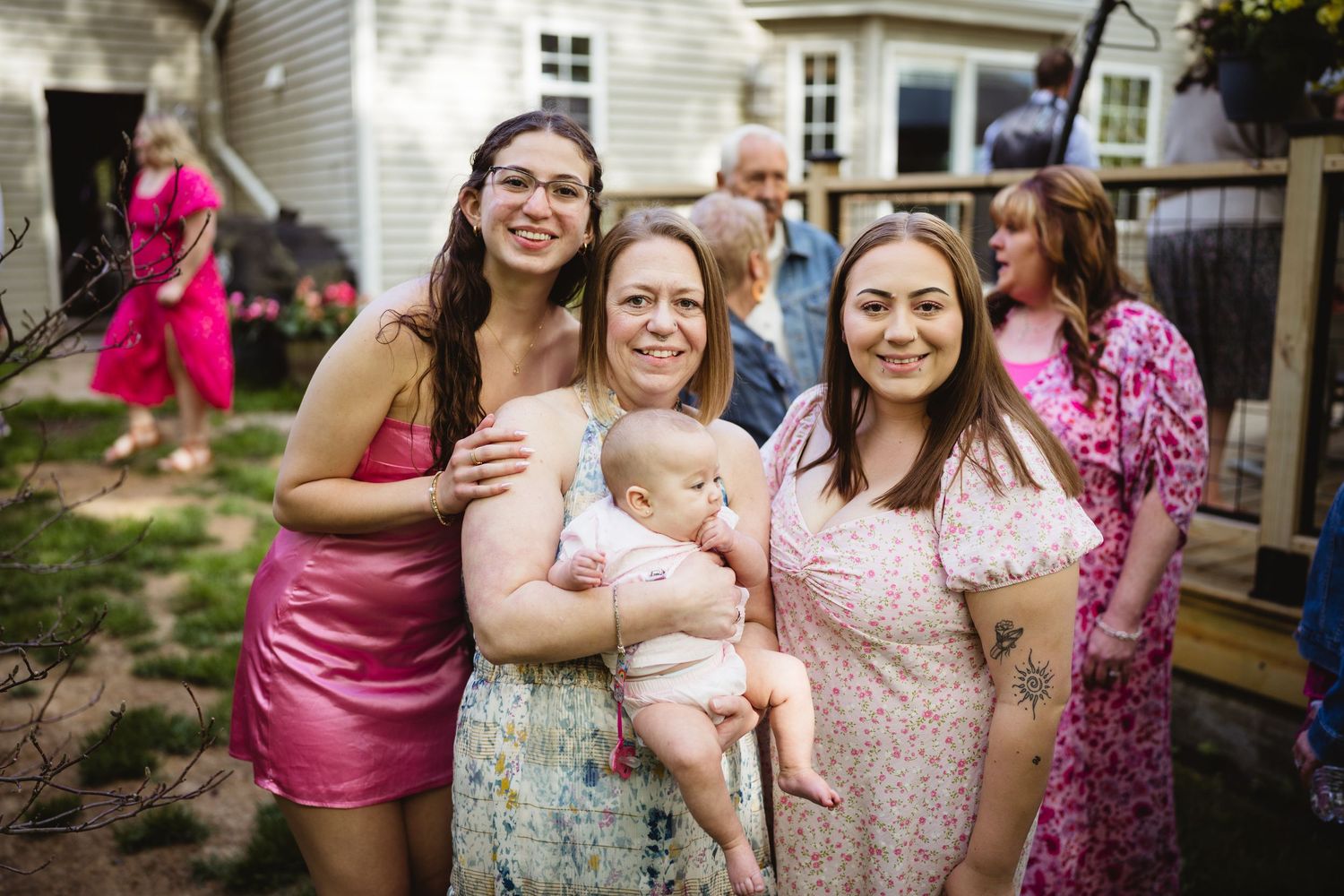 A group celebrates together at an outdoor gathering in a backyard, wearing pink and floral dresses while holding a baby.