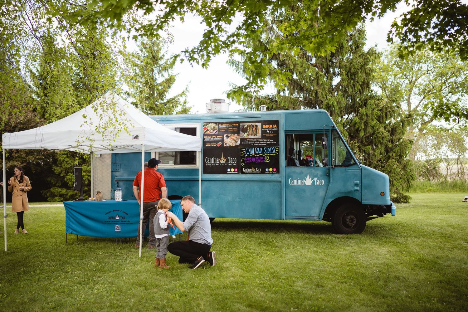 A blue food truck with a canopy tent setup serves customers on a grassy lawn under trees.