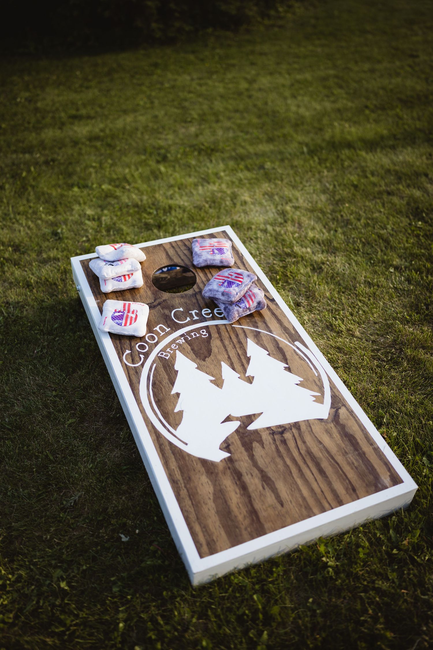 Wooden cornhole game board with pine tree design sits on grass with scattered bean bags on top.