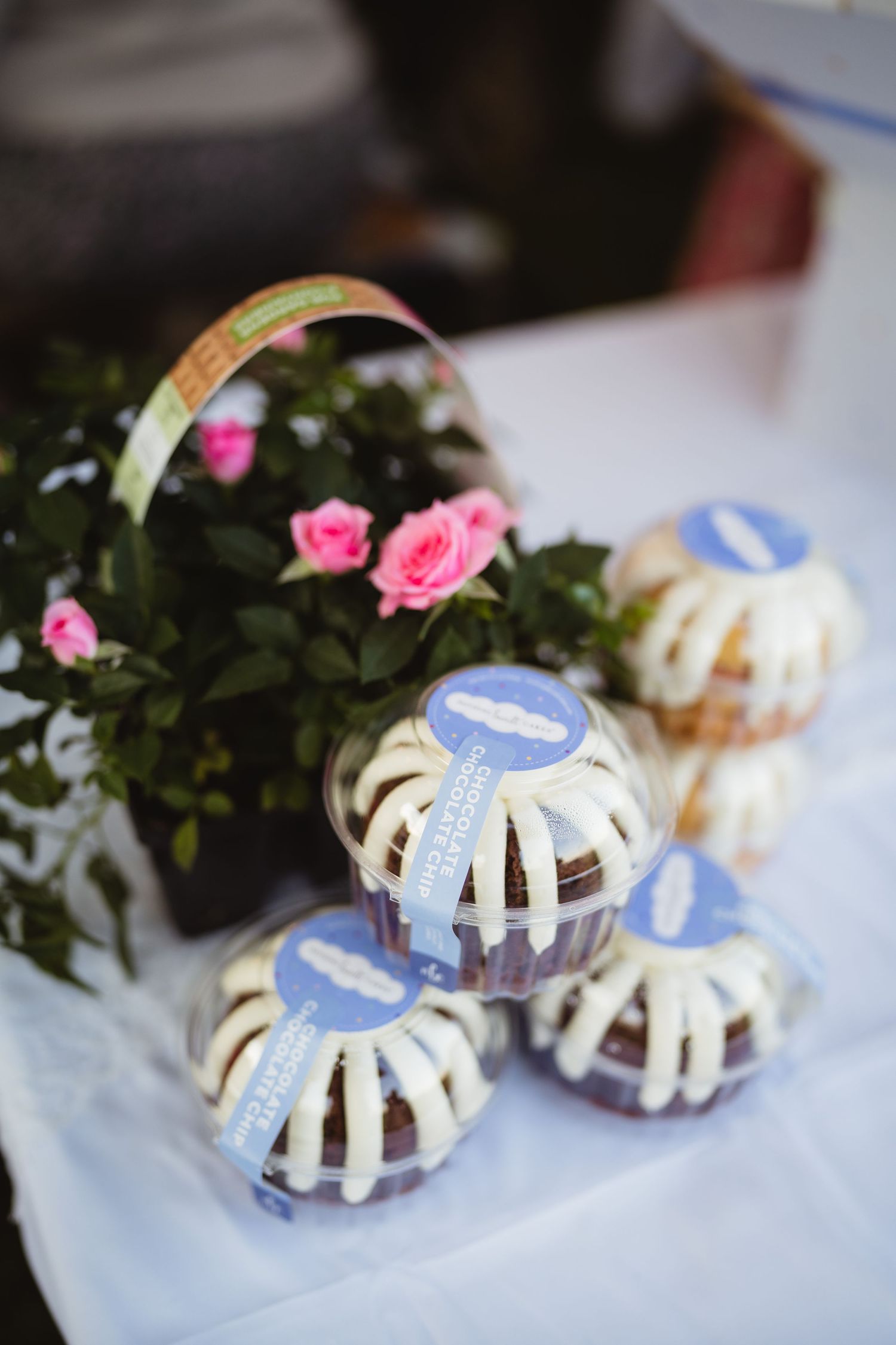 Miniature bundt cakes with white drizzle and blue labels displayed next to pink roses in a decorative arrangement.