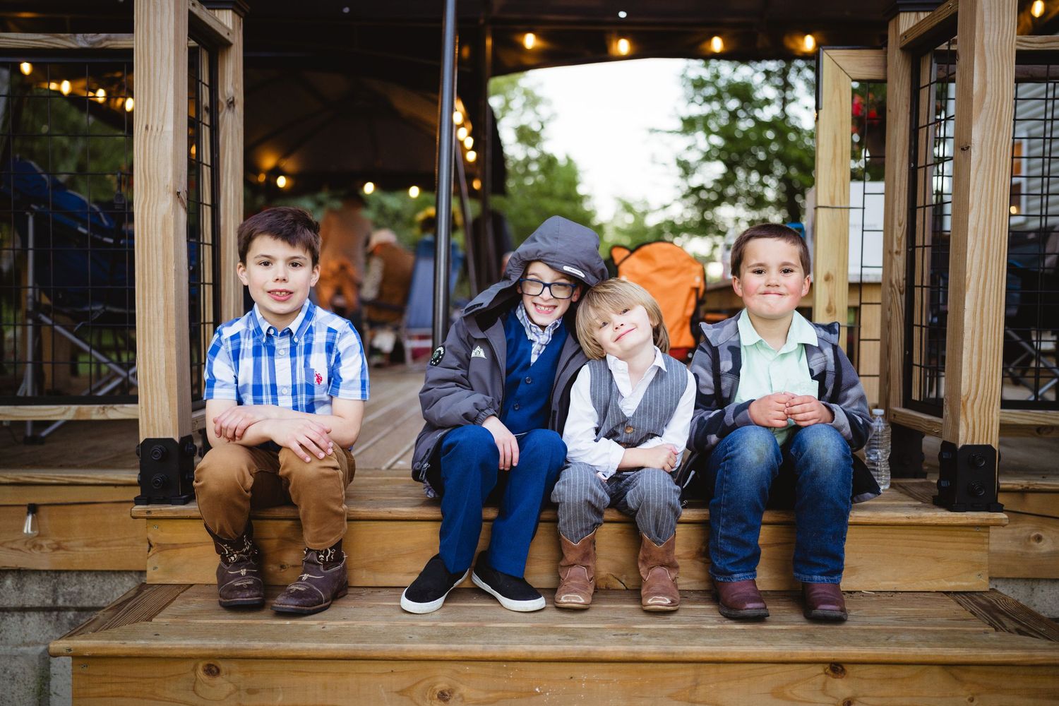 Four young boys sit together on a wooden porch wearing casual clothing.