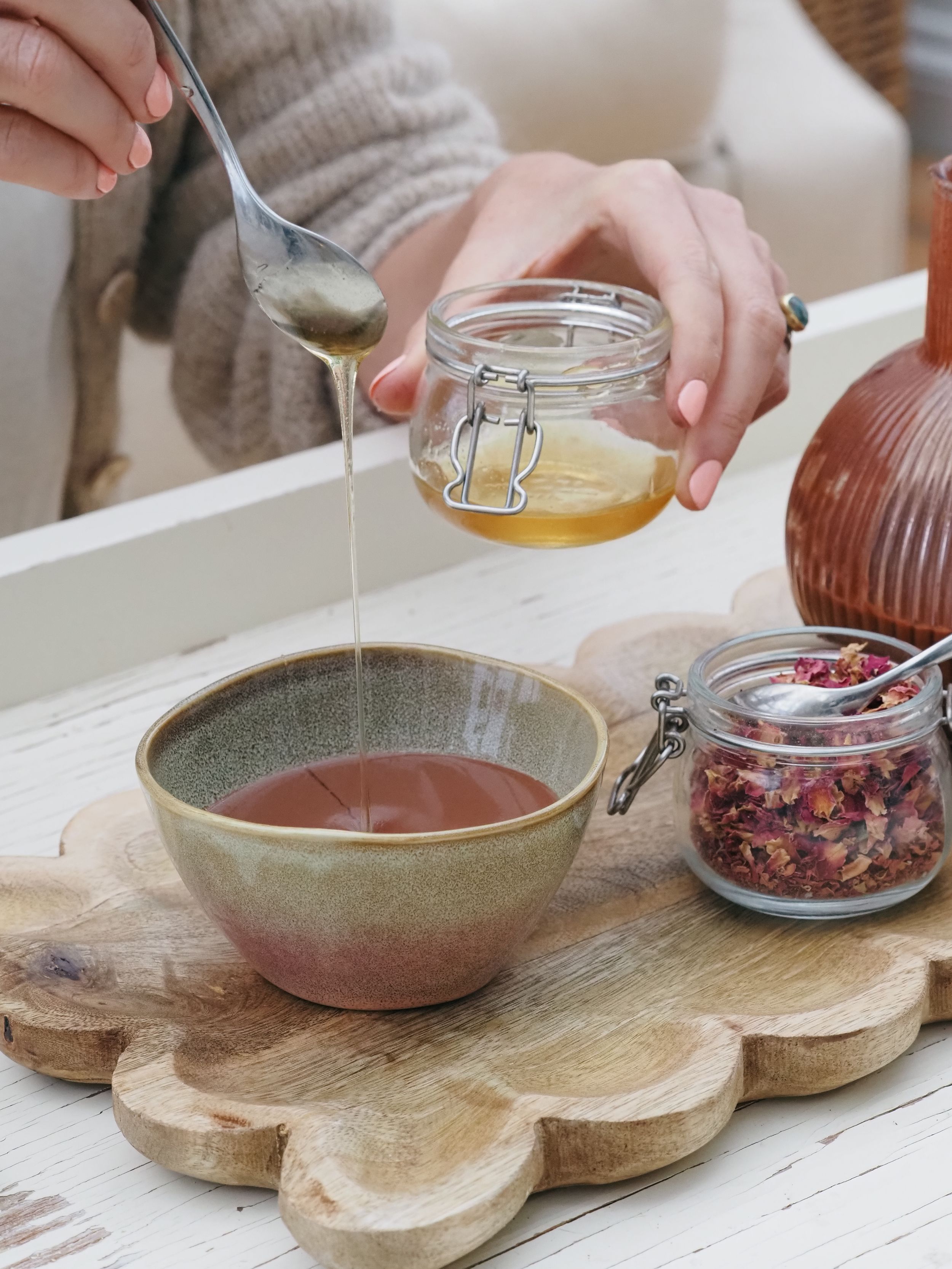 Hands drizzling honey into a ceramic bowl on rustic wooden board with a glass jar and rose petals preparation scene.