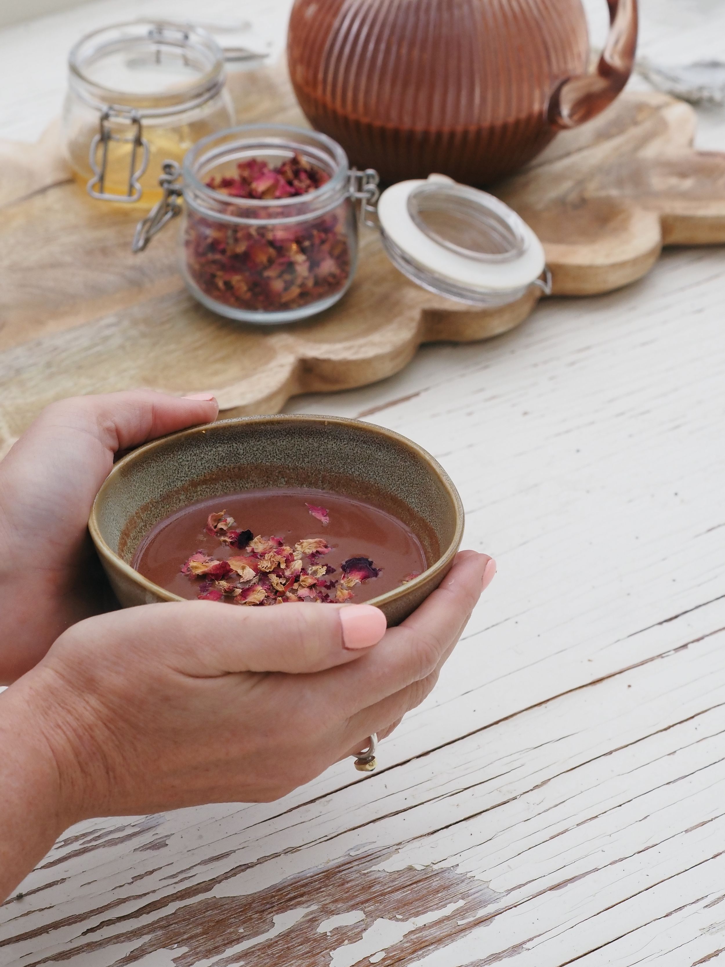 Hands holding a ceramic bowl of hot chocolate topped with dried goji berries next to a brown teapot on white wooden table.