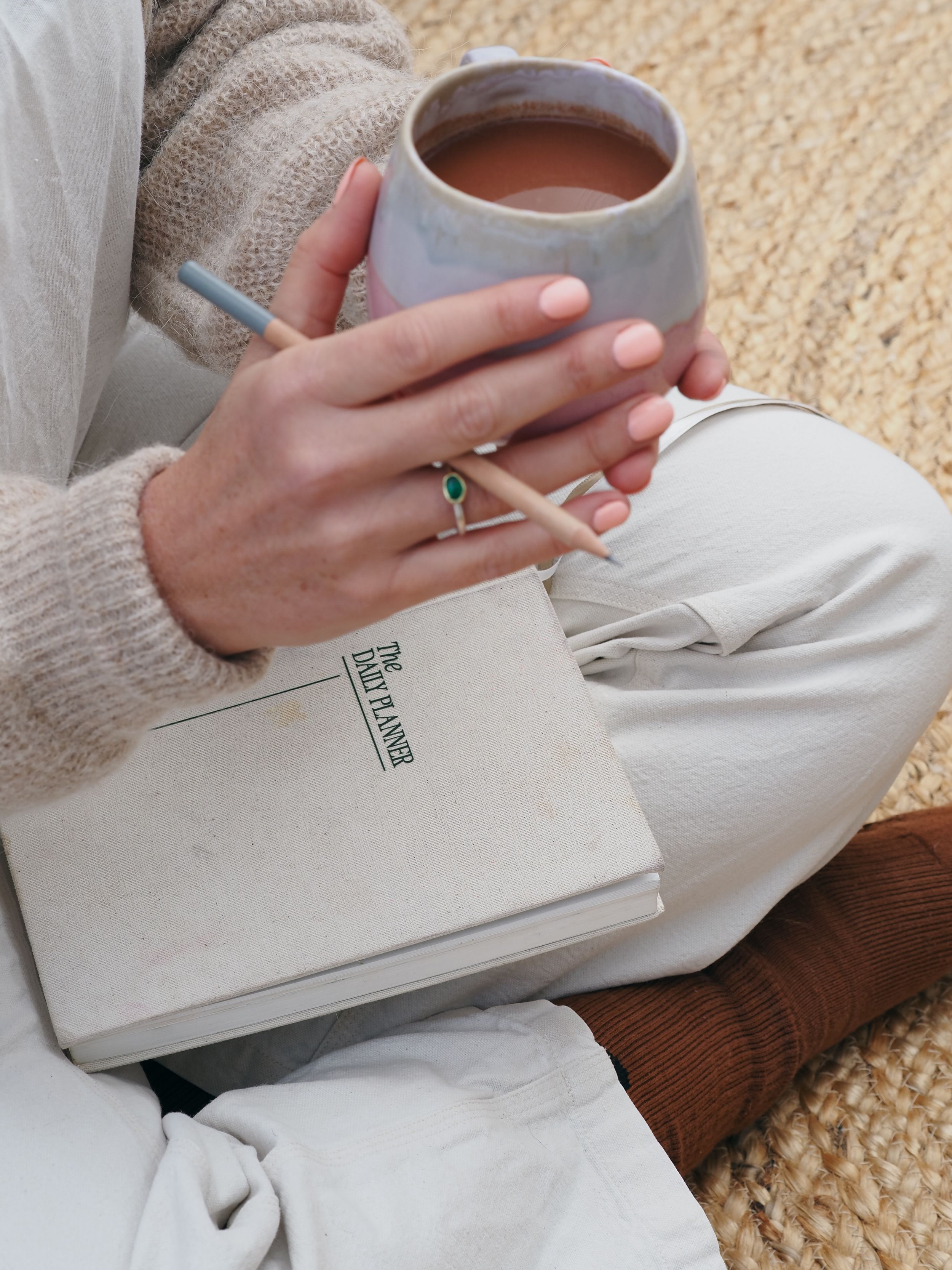 Hands holding a book and ceramic mug against white bedding and beige carpet background.