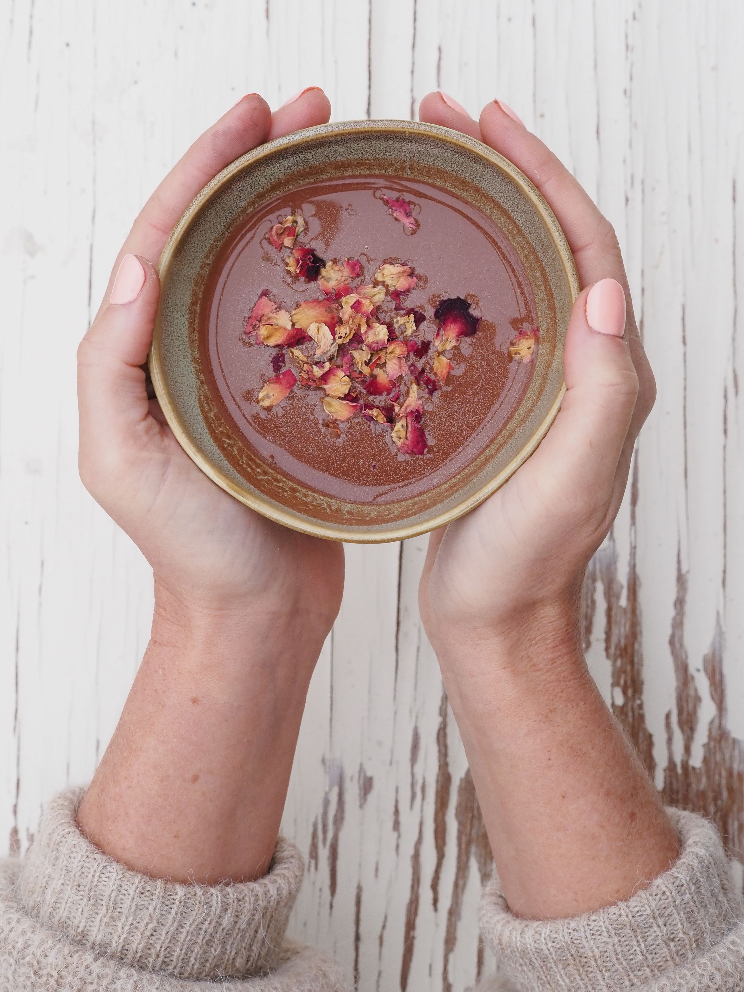 Hands holding ceramic bowls filled with chocolate pudding garnished with pink rose petals on a rustic white wood surface.