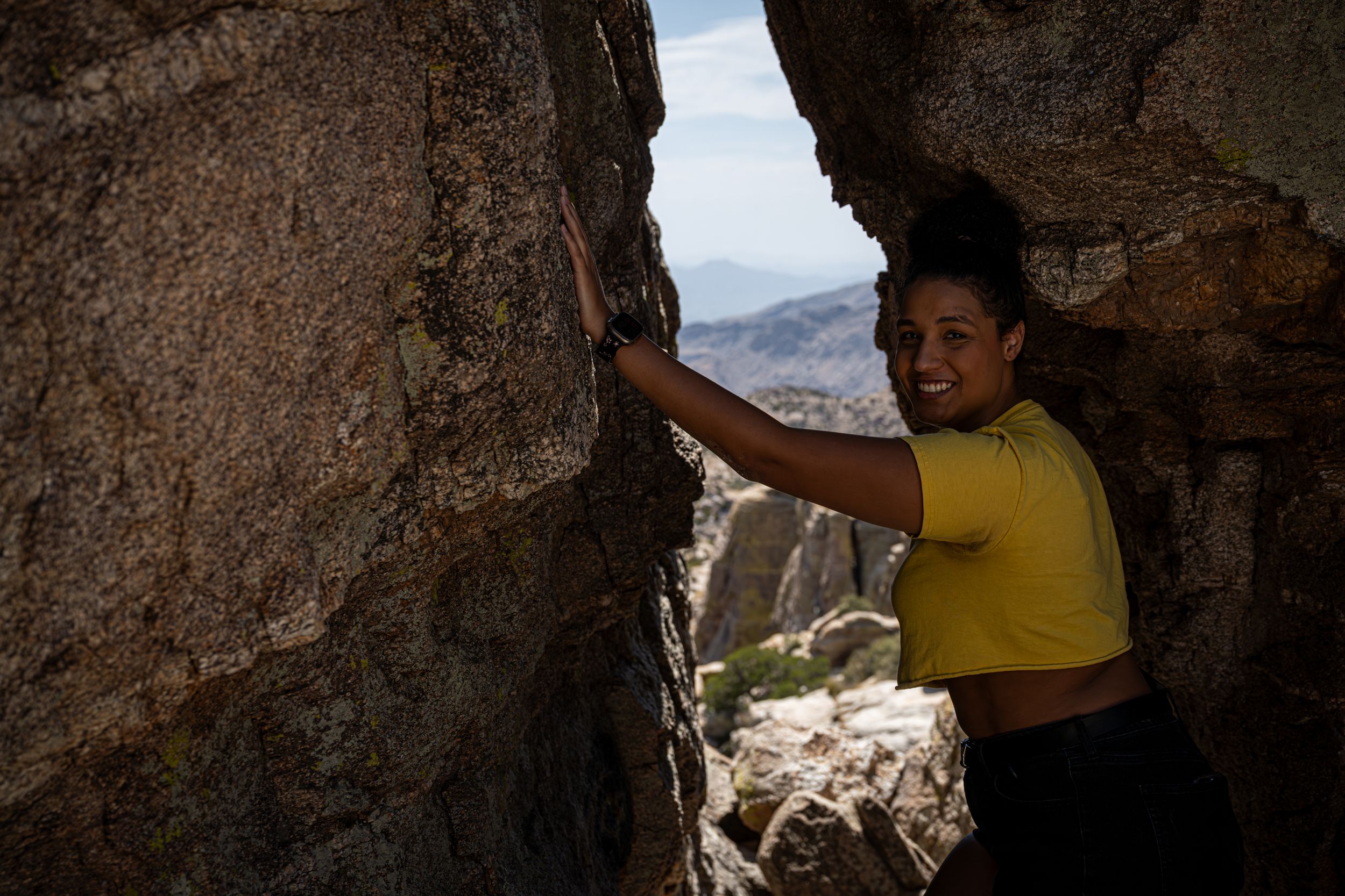 Person in yellow shirt exploring natural cave opening with mountain vista beyond.