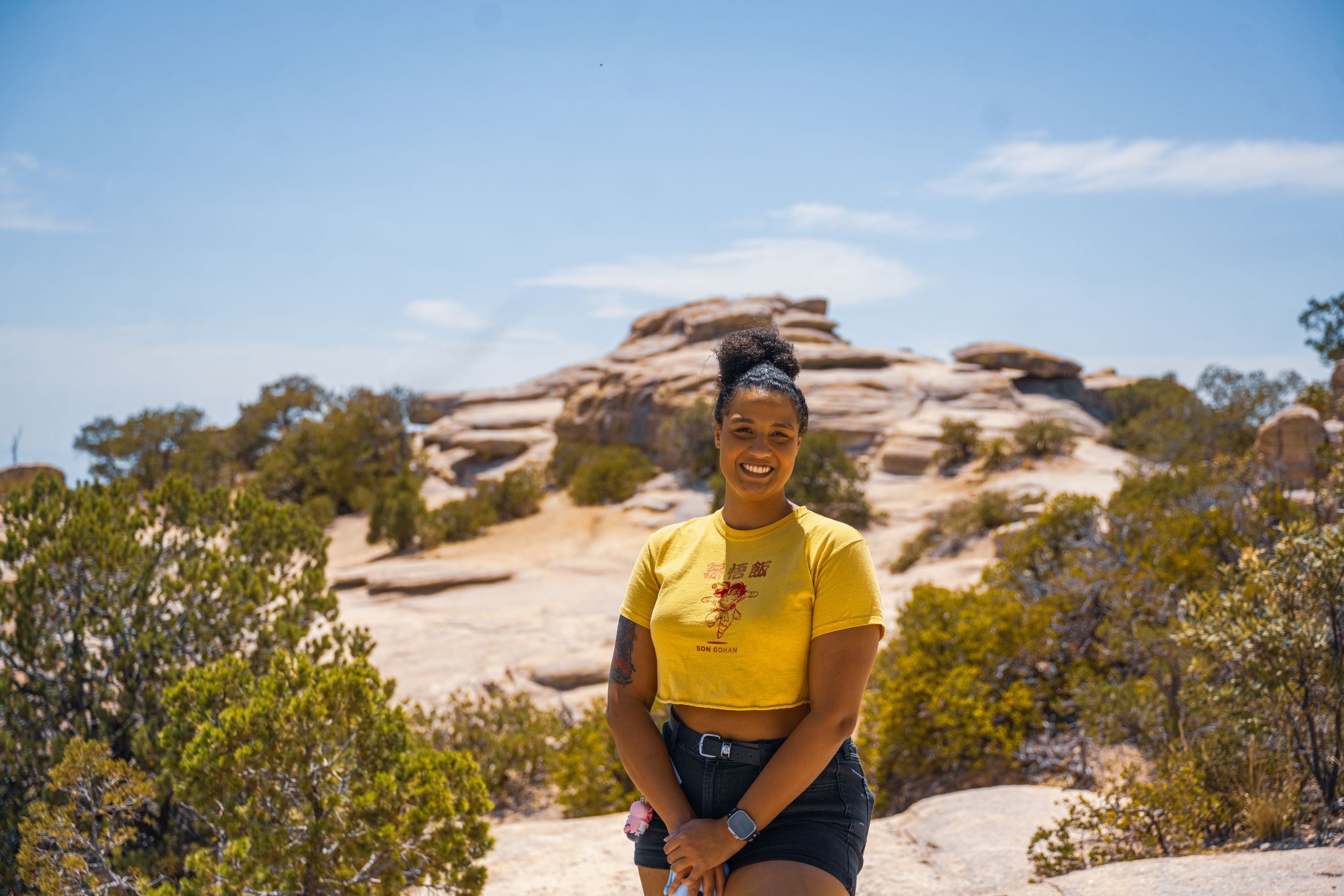 Person in yellow shirt walking along desert trail with rock formations and blue sky.