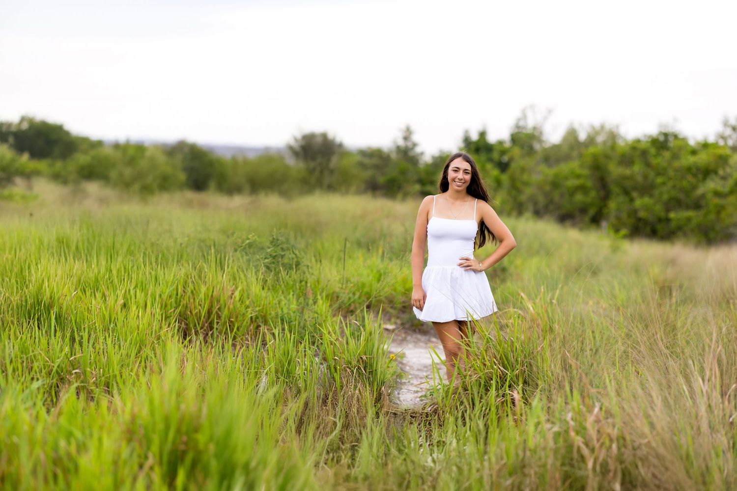 Woman in white sundress standing among tall grass in natural outdoor setting.