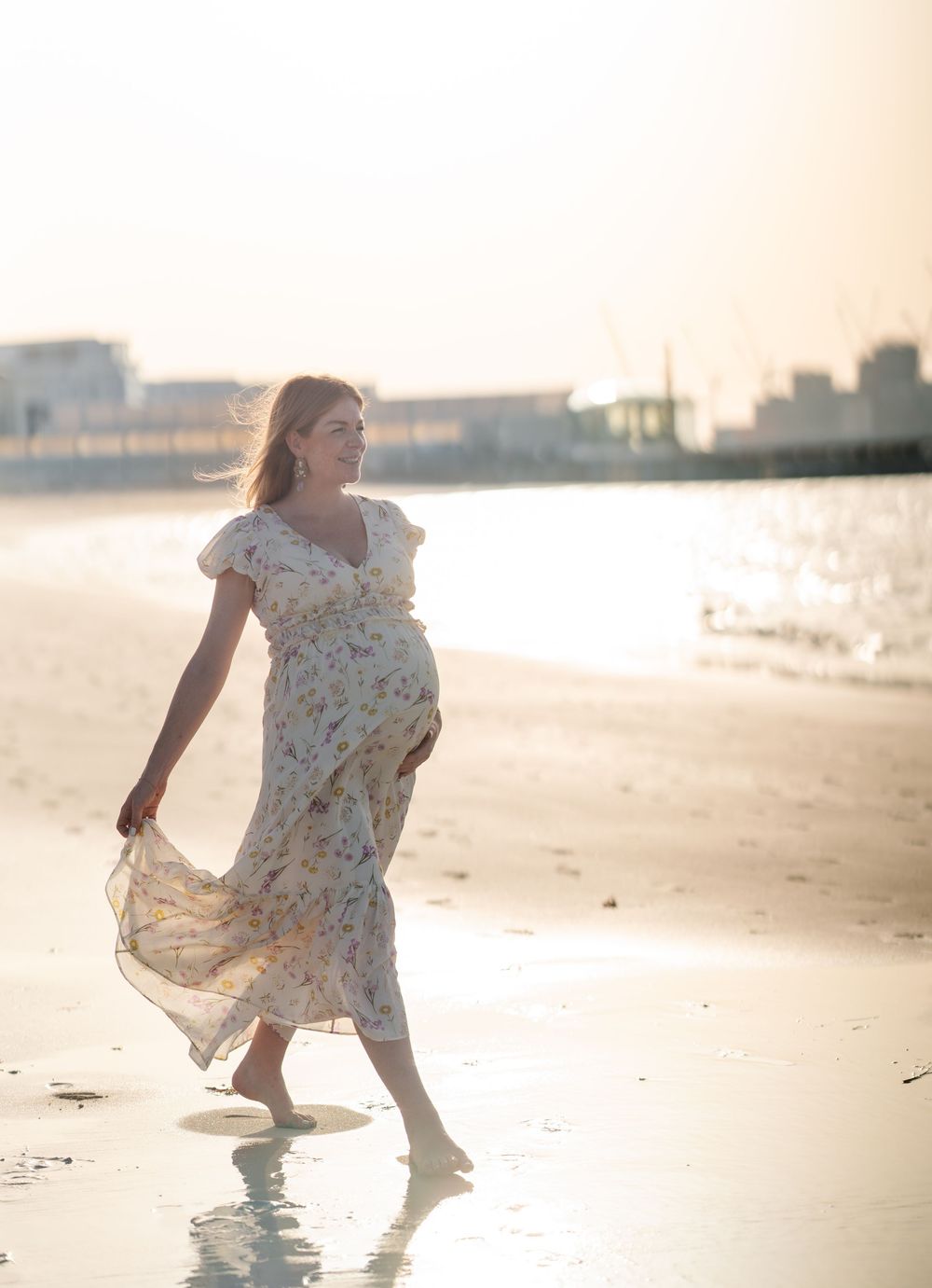 A pregnant woman in a floral dress walks along a beach at sunset.