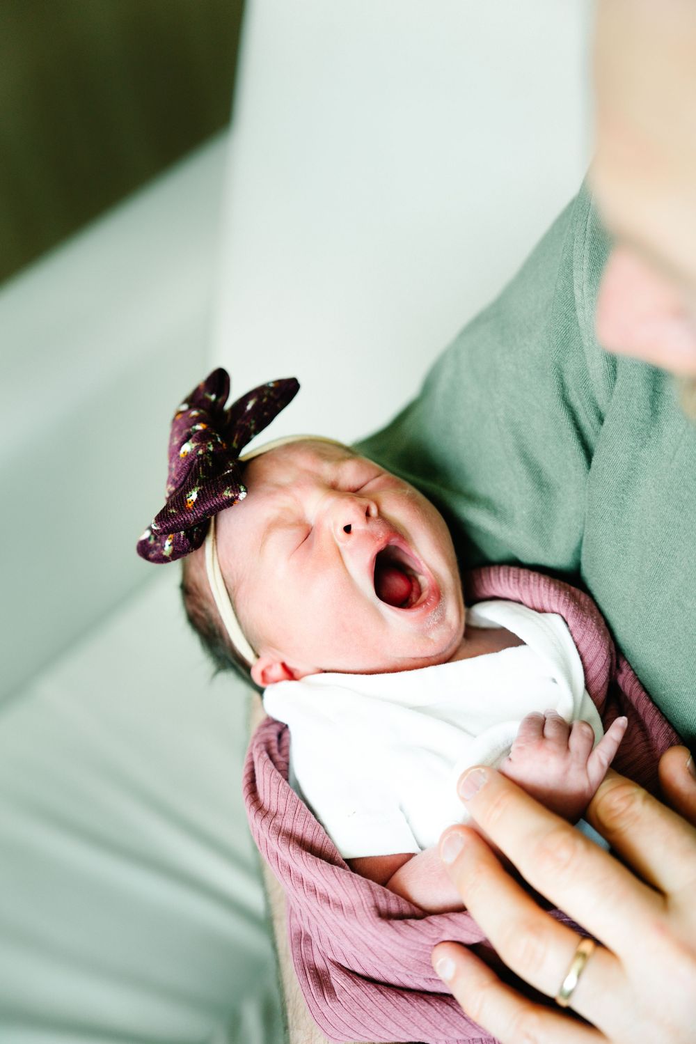 Newborn baby with butterfly hair bow yawns while being cradled in gentle arms.