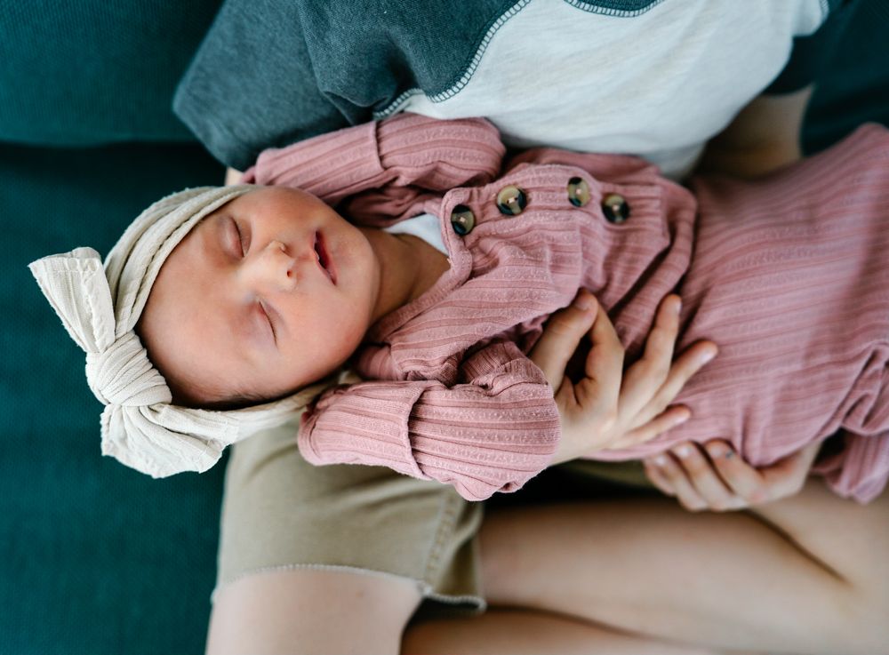 Peaceful newborn wearing cream bow and pink outfit rests on teal surface.