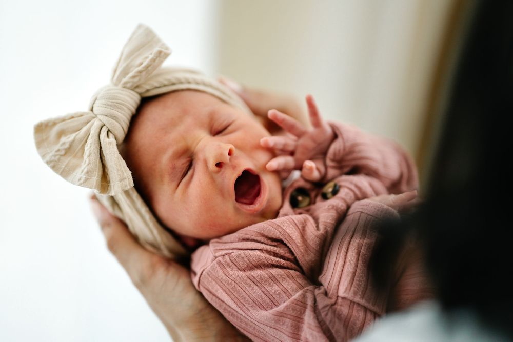 Sleepy newborn in mauve outfit and beige bow headband yawns sweetly.
