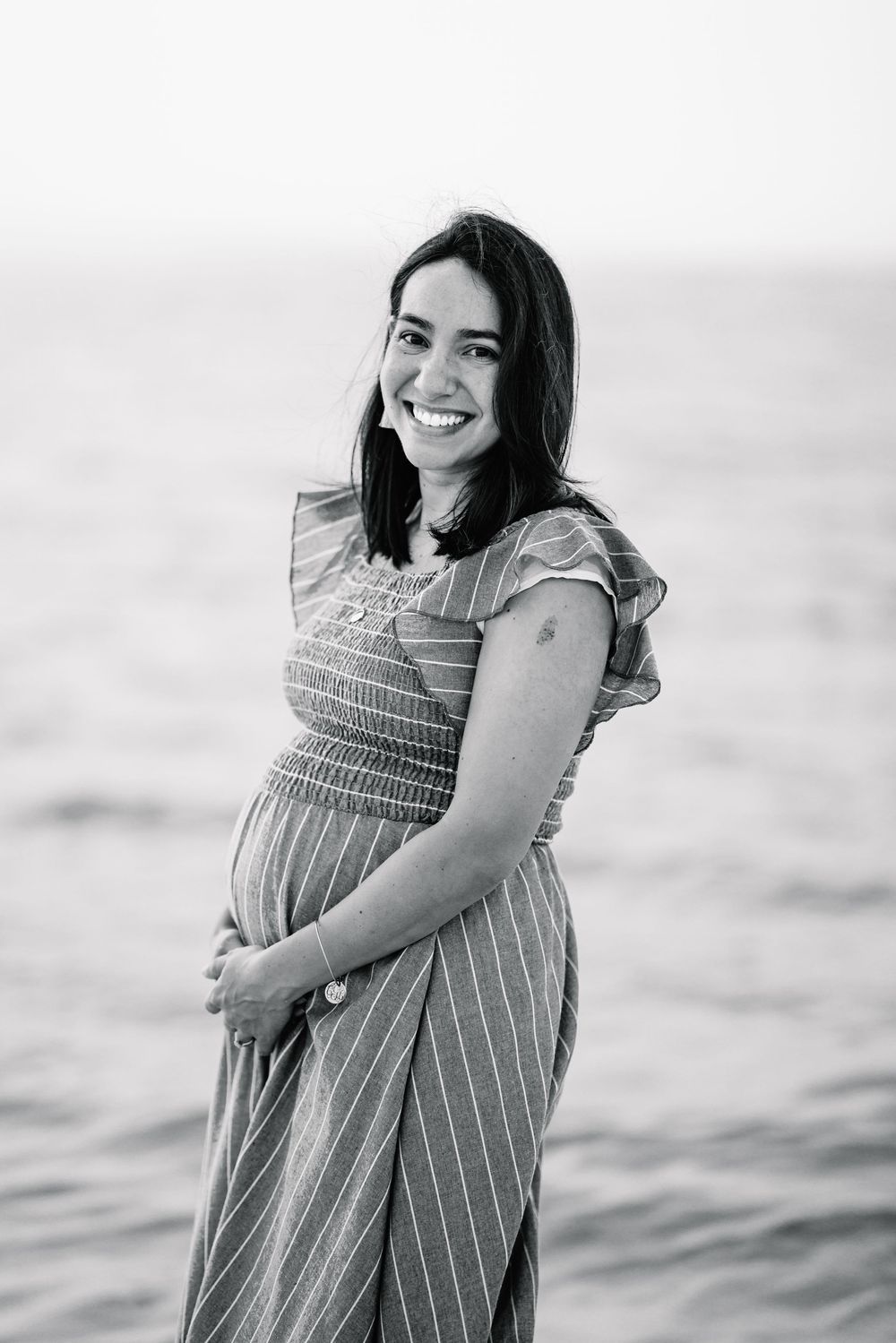 Expectant mother in striped dress smiles at beach in black and white photo.