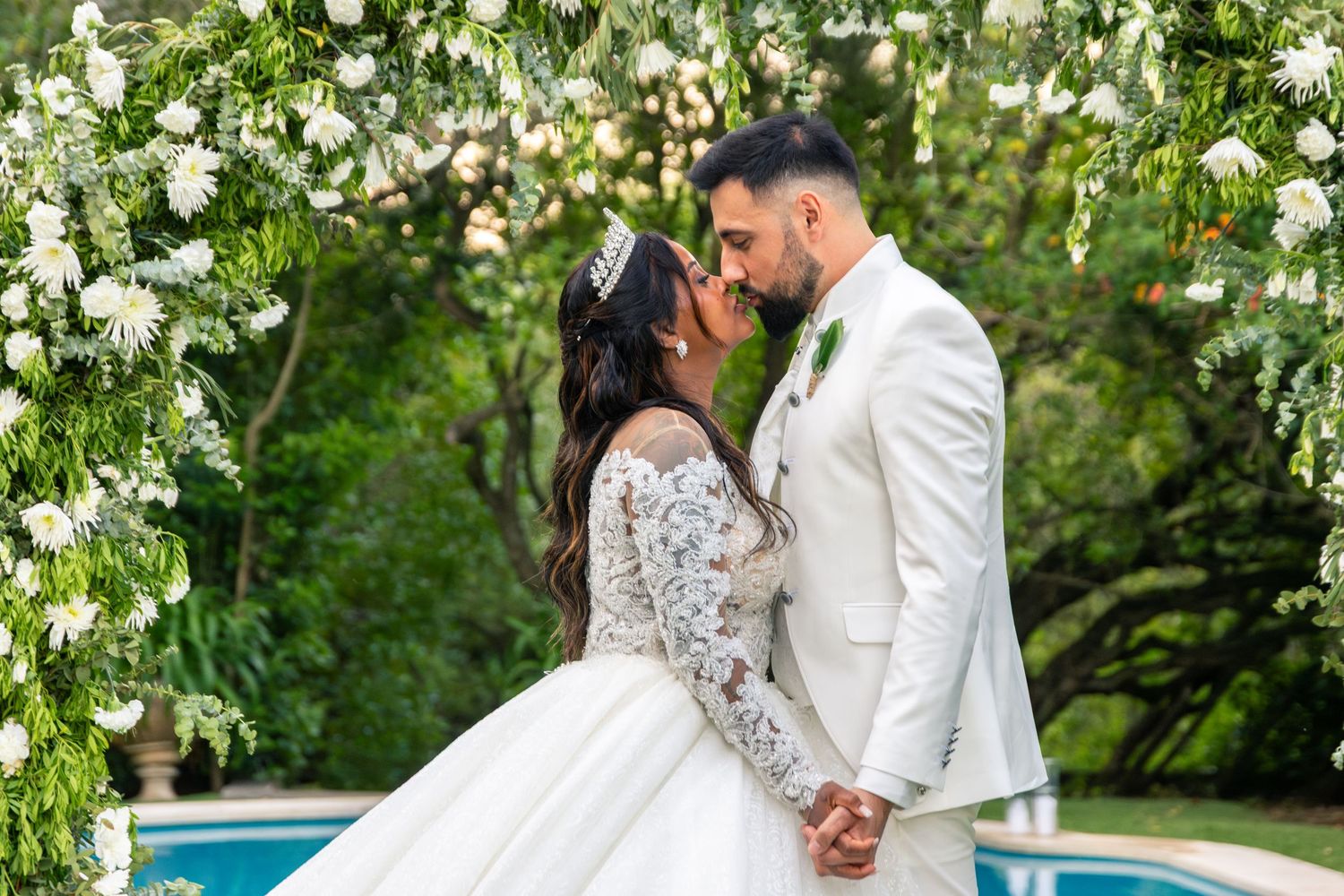 Romantic wedding couple embracing under white floral arch near pool in garden setting.
