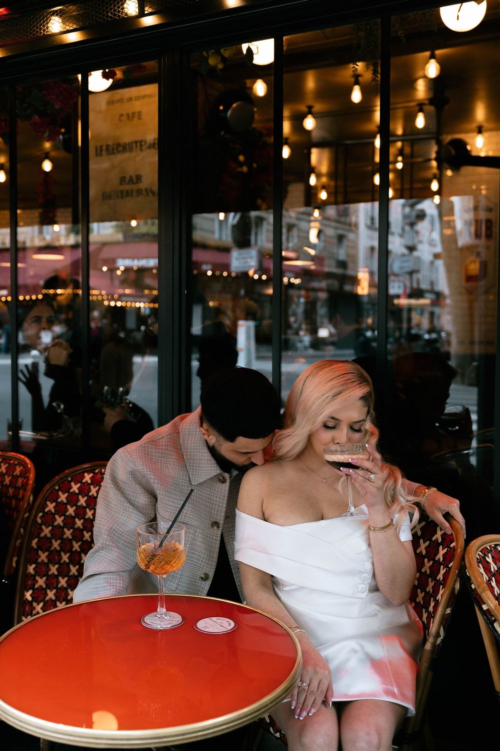 Couple enjoying drinks at a romantic Parisian sidewalk cafe with string lights and red table in warm evening ambiance.