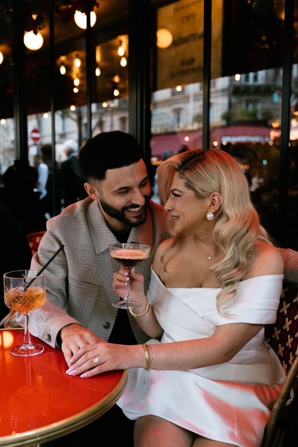 Couple enjoying cocktails and sharing romantic moment at illuminated Parisian cafe with red table.