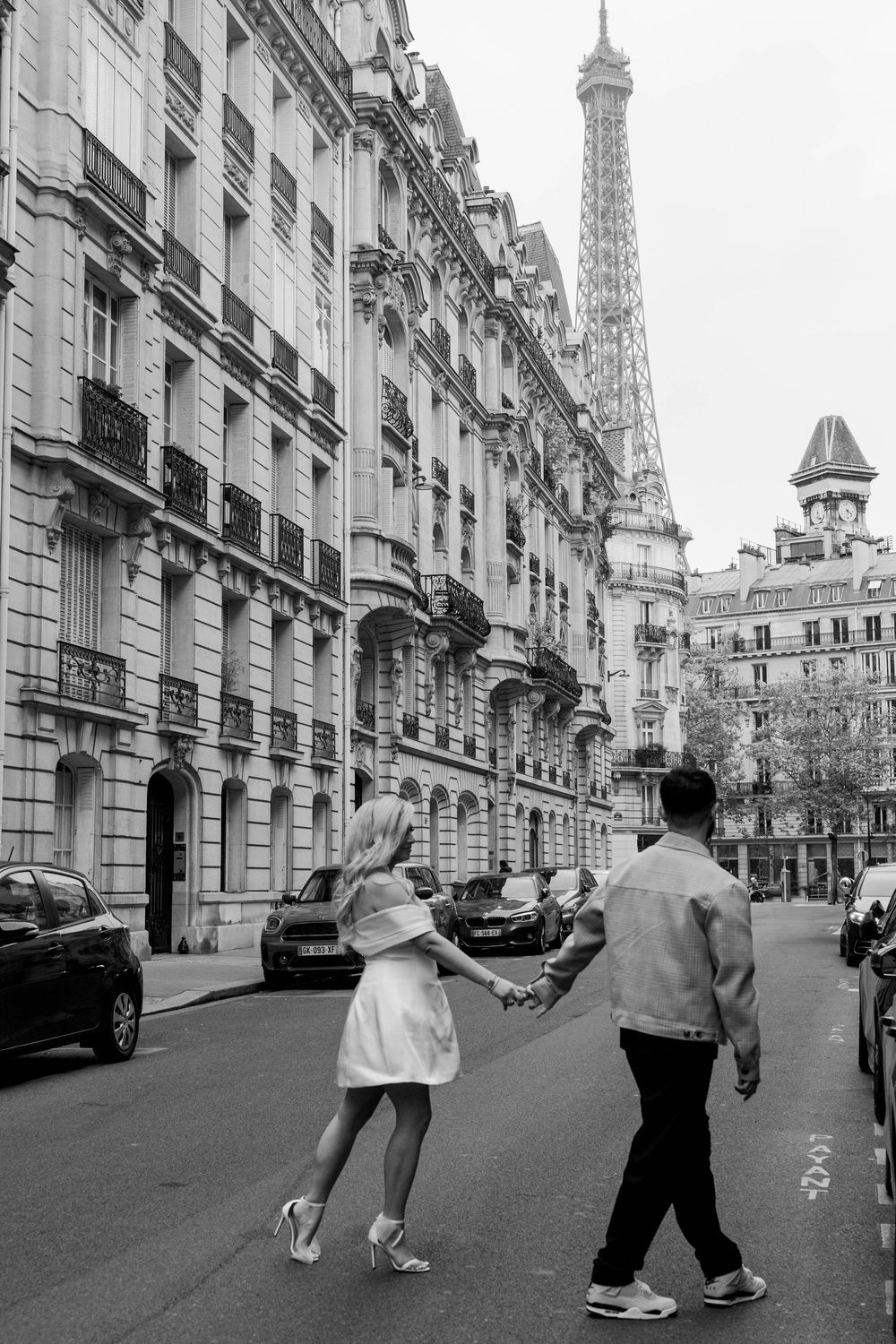 Couple walking down a romantic Parisian street with iconic Eiffel Tower visible in the black and white cityscape.
