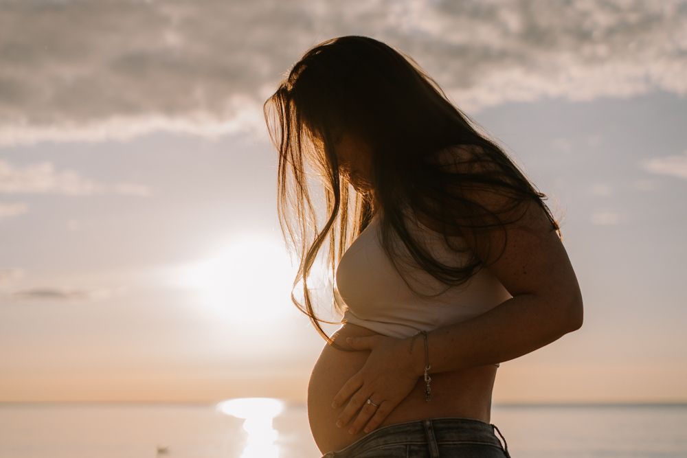 PREGNANT WOMAN AT THE BEACH ON A SUNRISE, LOOKING AT HER BELLY