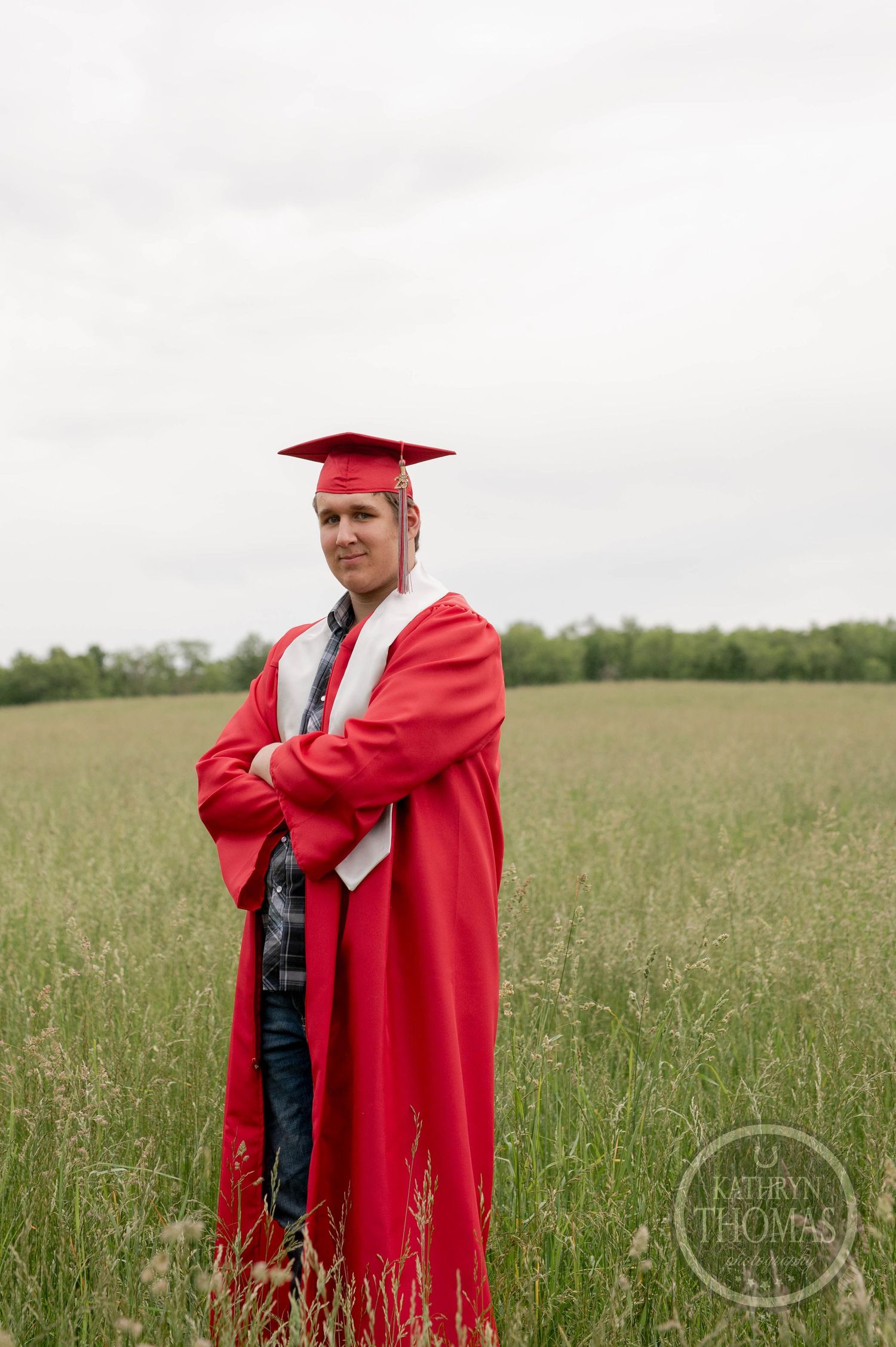 Graduate in red cap and gown stands confidently in a grassy field during outdoor graduation photo shoot.