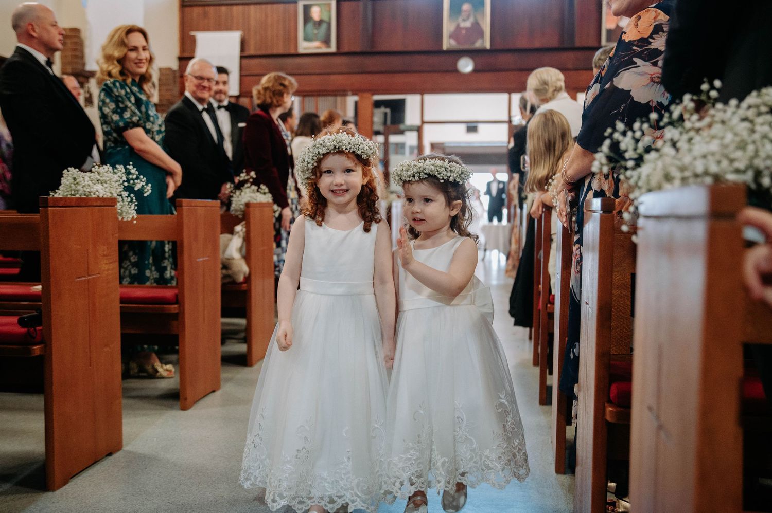 Two young flower girls in white lacy dresses walk down the church aisle decorated with baby's breath flowers.