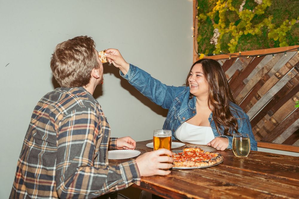 Two people enjoying craft beer and pizza while sitting at a rustic wooden table in a casual dining setting.