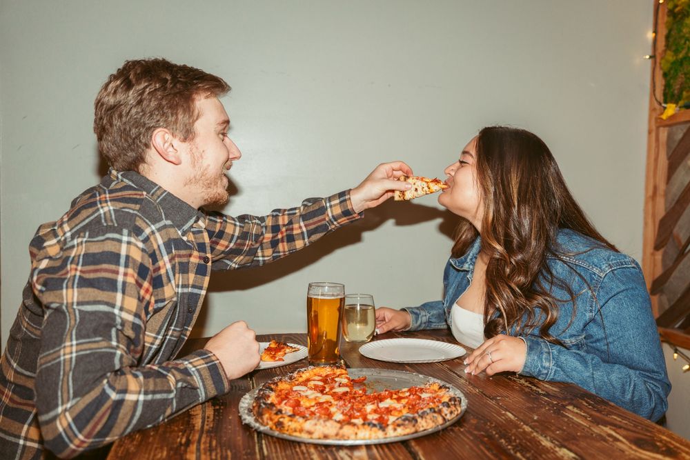 Two people share a pizza and beer at a wooden table while enjoying a casual dining experience together.