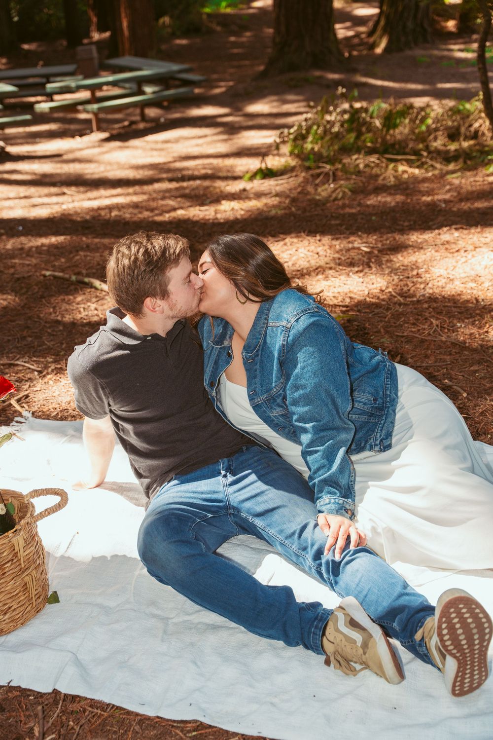 Romantic couple sharing a kiss while sitting on a white blanket during a picnic in a wooded area.