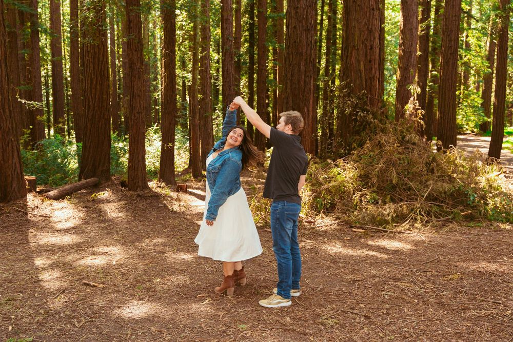 A couple dancing playfully on a dirt path surrounded by tall redwood trees in a sunlit forest.