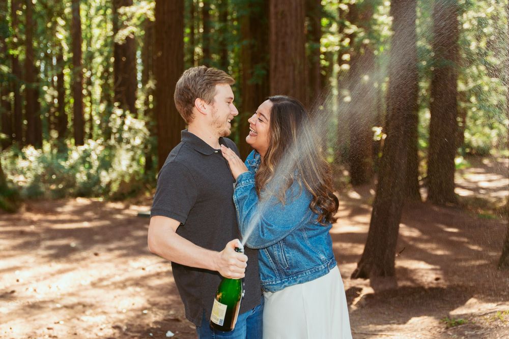 A romantic couple embraces among tall redwood trees in a sunlit forest setting.
