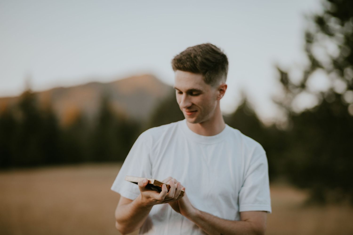 Someone in a white t-shirt checks their phone outdoors with mountains and trees in the background at dusk.