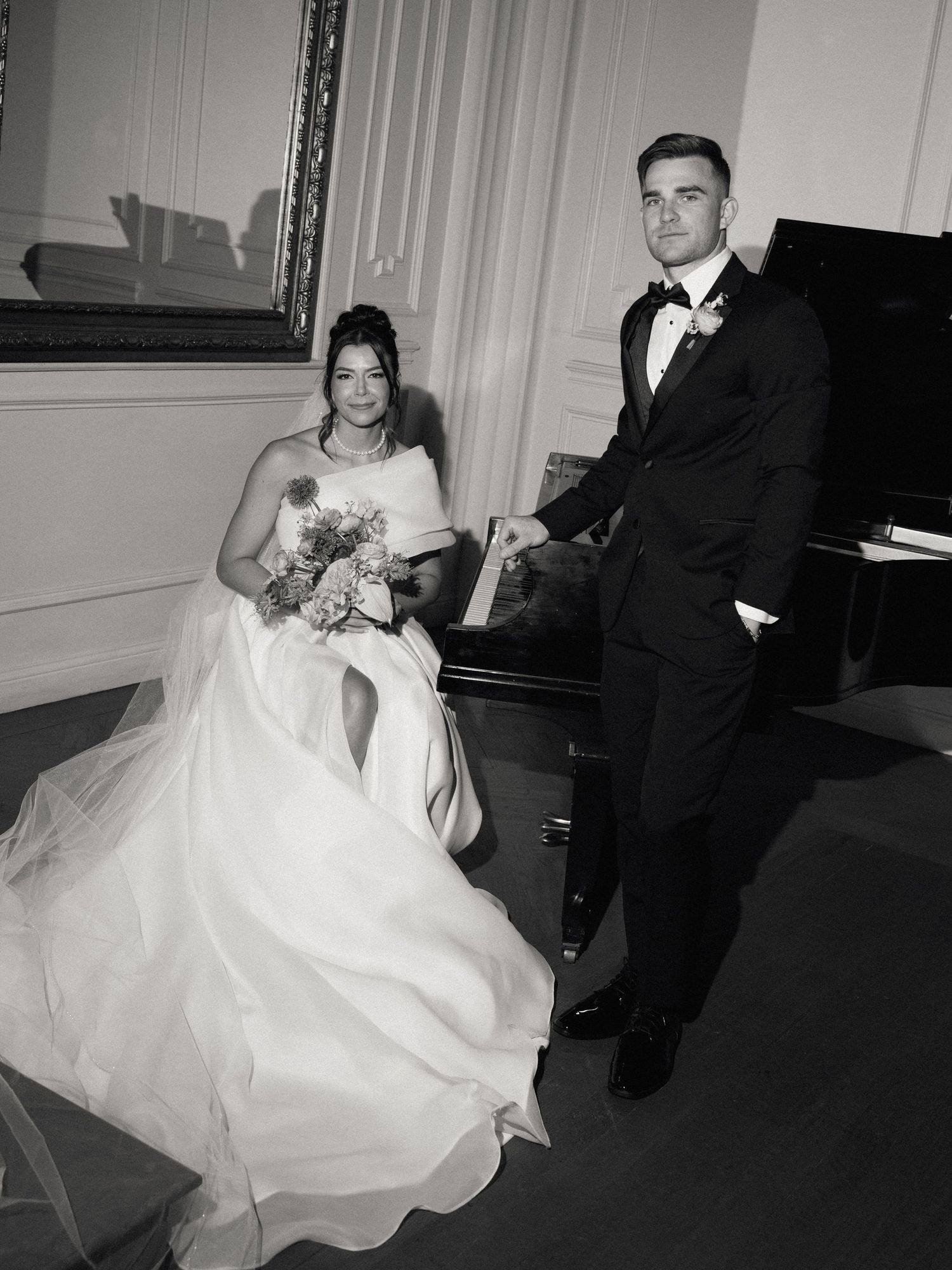 Newlywed couple poses elegantly by a grand piano in a formal wedding setting with traditional attire and colorful bouquet.