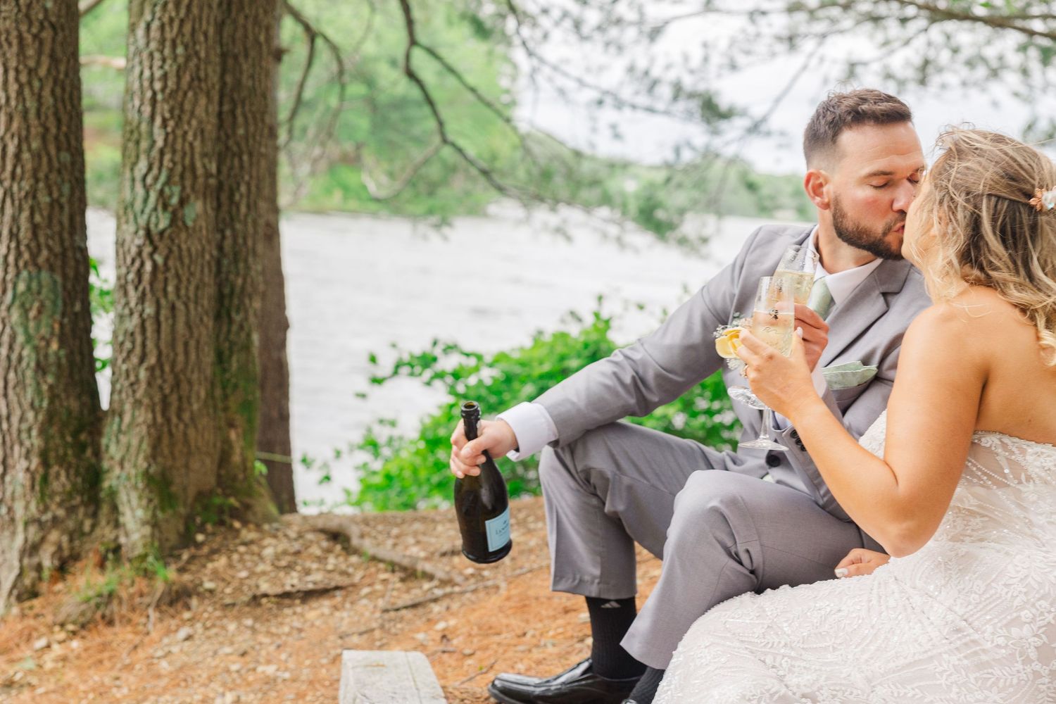 A couple in formal attire sits together by a lake enjoying champagne beneath tall trees on a romantic afternoon.