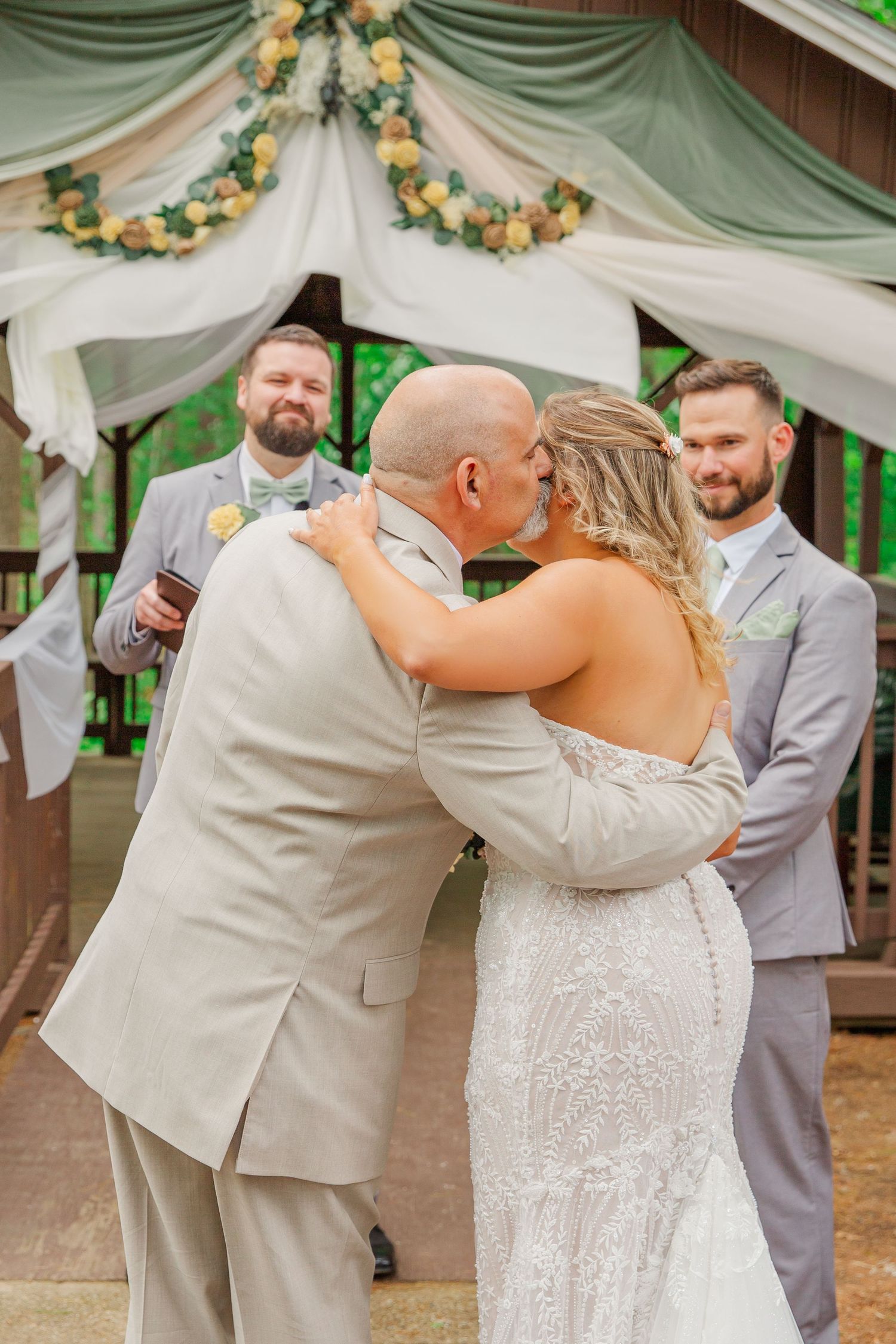 A wedding ceremony moment under an elegant white and sage green draped gazebo adorned with yellow floral garlands.