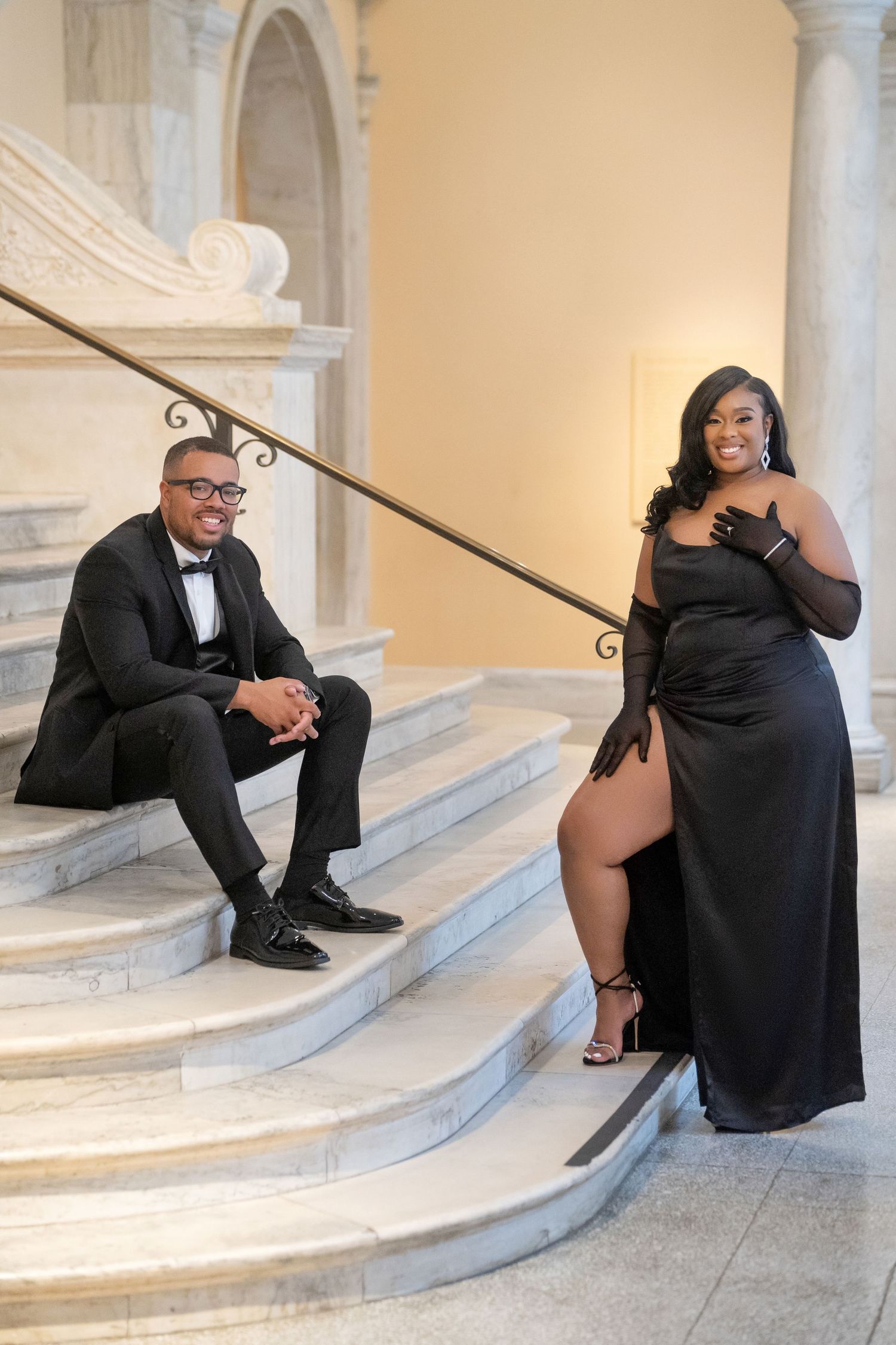 Elegant formal couple pose on grand marble staircase with ornate railing in formal black evening attire.