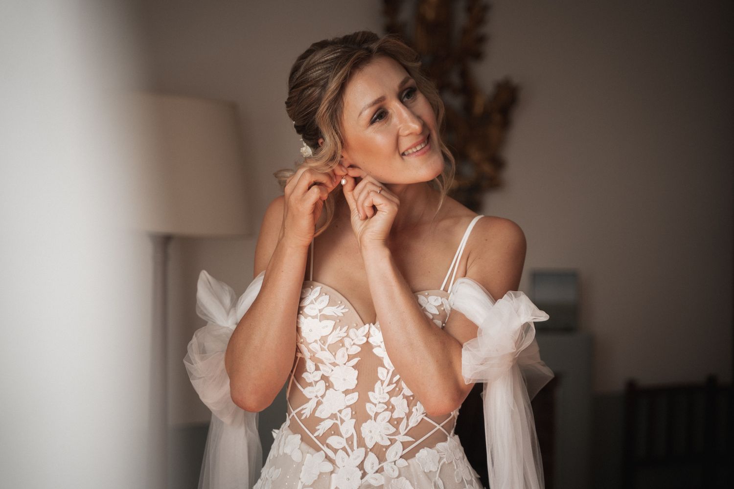 A bride in an elegant lace wedding gown with sheer details and puffed sleeves smiles while putting on earrings.
