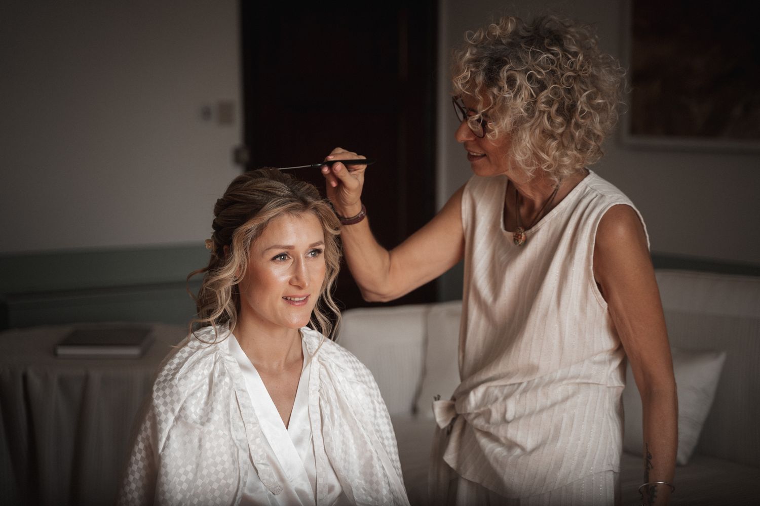 A hairstylist works on a client's hair in a soft, moody setting with warm lighting.