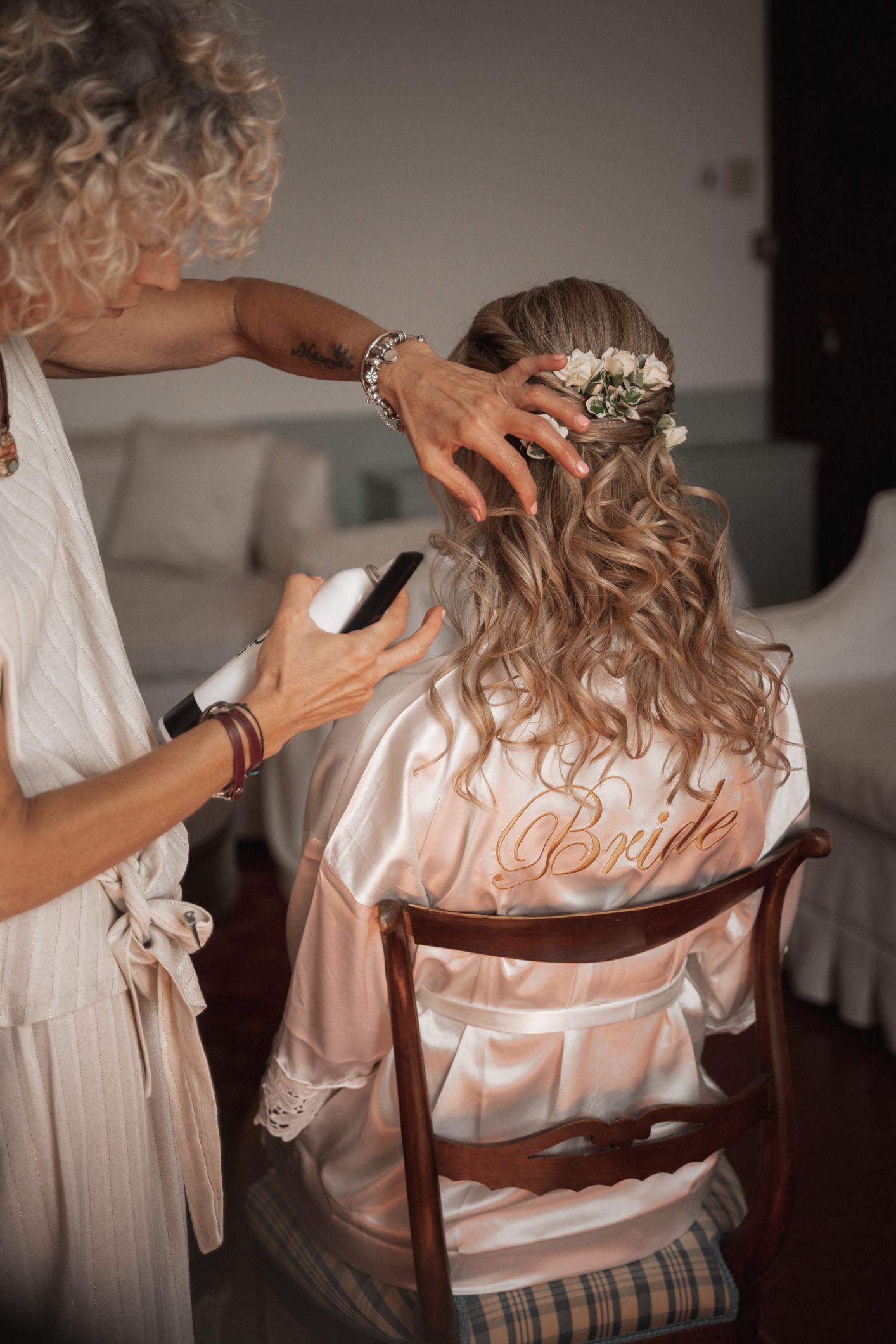 Hairstylist arranges delicate flowers in a bride's wavy hair while she wears a pink satin robe during wedding preparations.