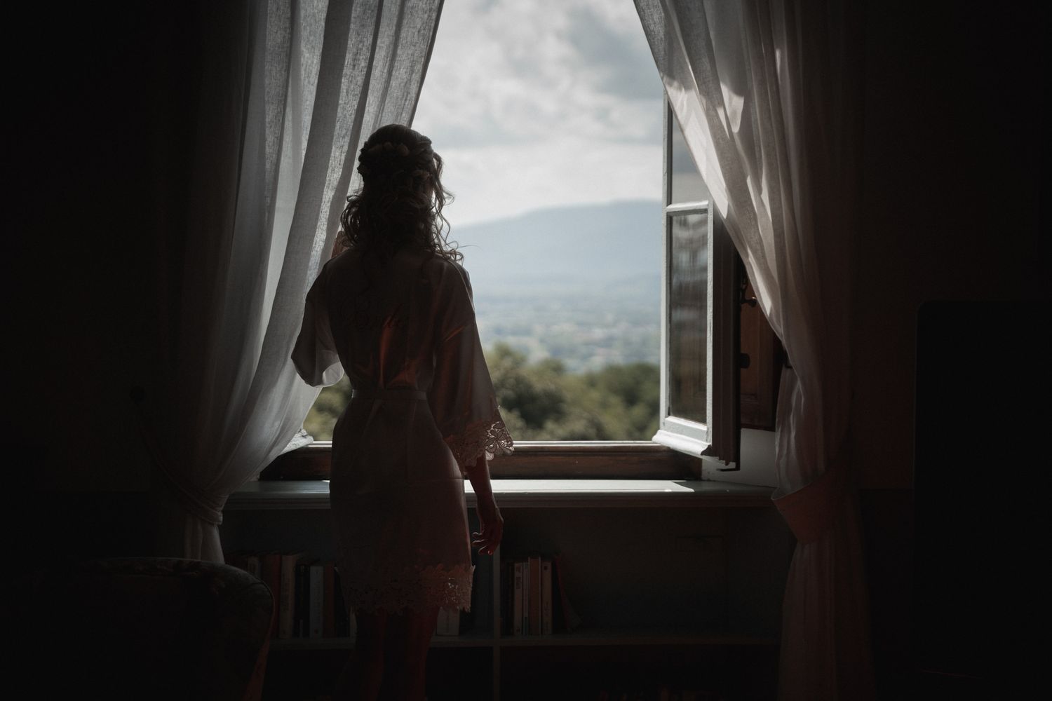 A silhouetted figure in a robe stands at a window looking out at mountain views through white curtains.