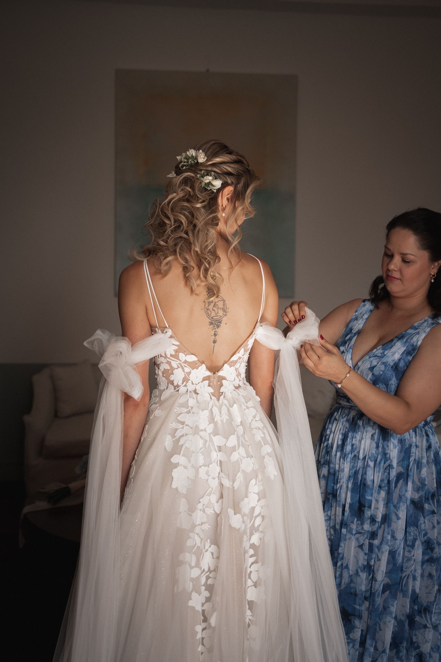 A bride in a white floral wedding gown with low-back detail gets help adjusting her dress from an attendant in blue.