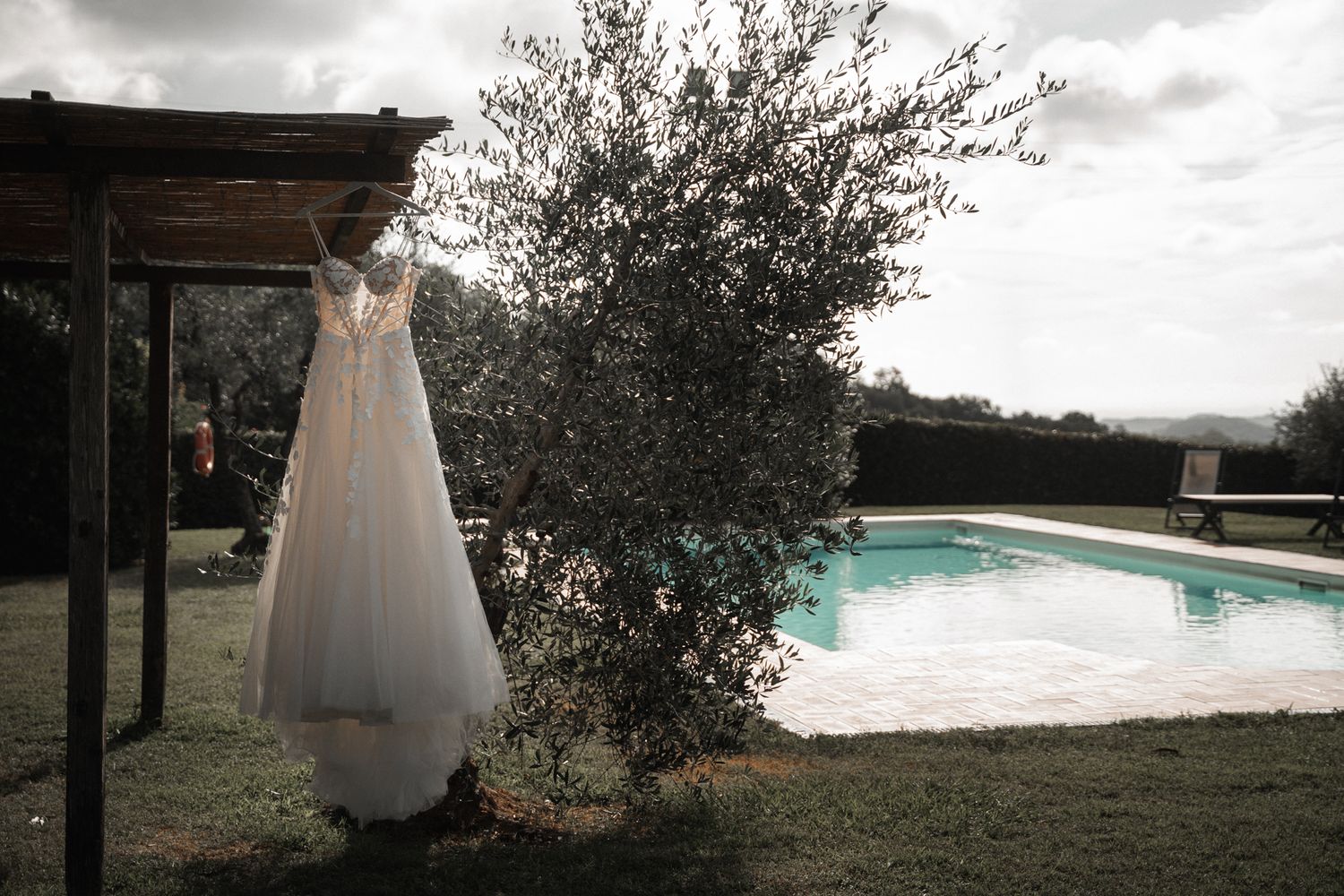 White wedding dress hanging from pergola beside swimming pool with olive tree under cloudy sky.