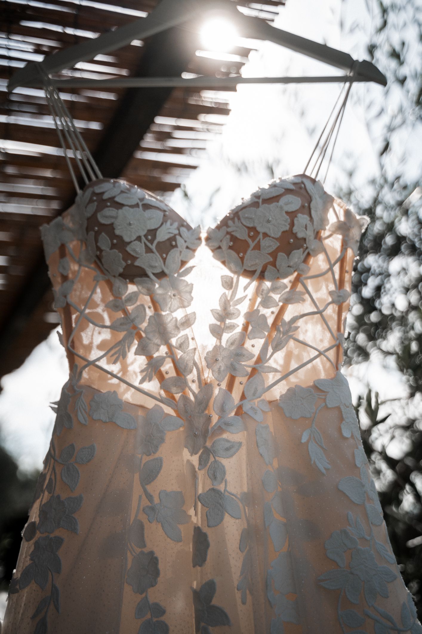 Wedding dress with intricate floral lace detail hangs in sunlight against green bokeh background creating ethereal glow.