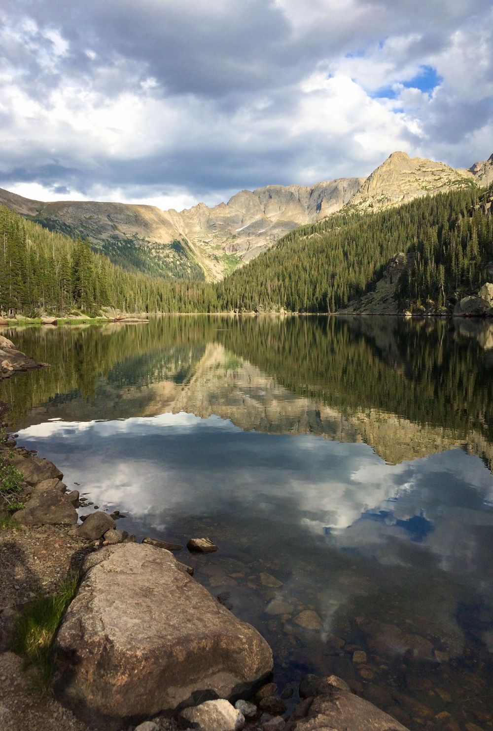 Mountain lake reflecting evergreen forest and peaks under cloudy skies.