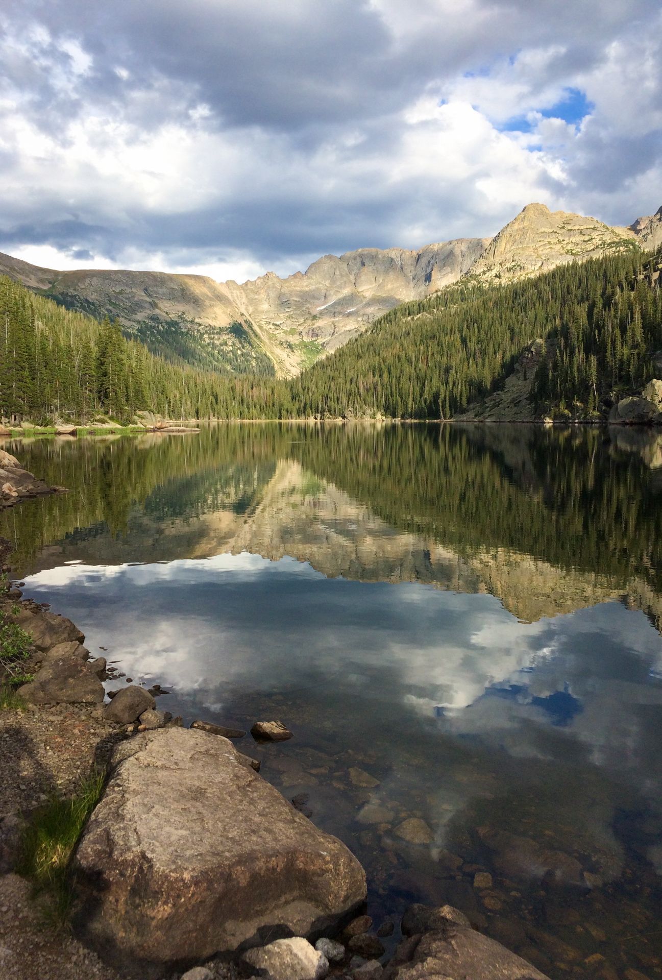 Mountain lake reflecting evergreen forest and peaks under cloudy skies.