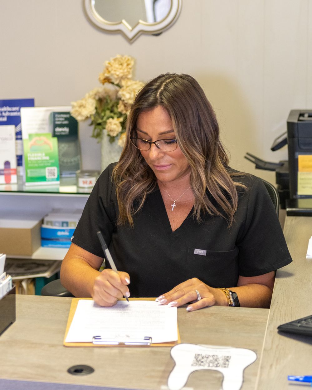 Healthcare worker in black scrubs completing paperwork at a medical desk with supplies visible in background.