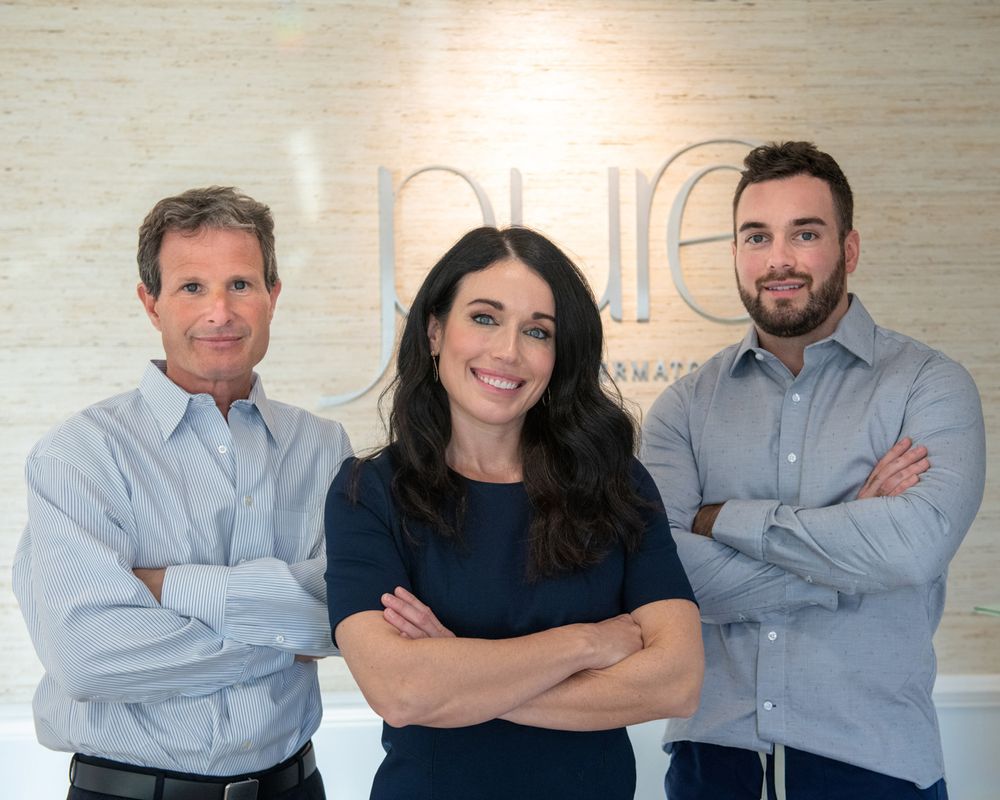 Three team members pose in office with arms crossed in front of logo wall.
