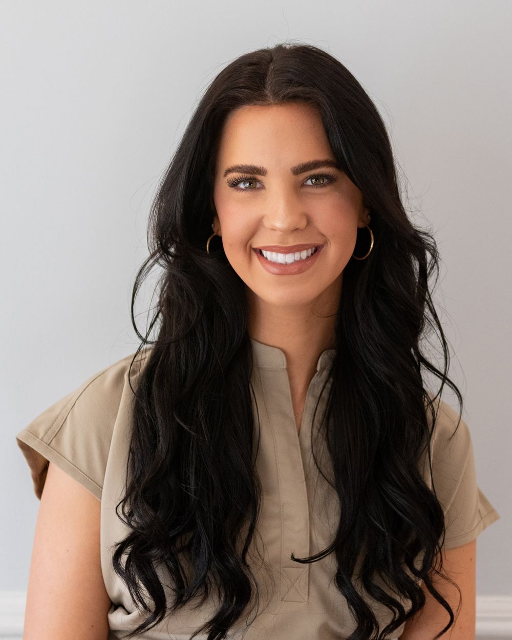 Healthcare provider wearing beige scrubs smiling against grey background in professional headshot.