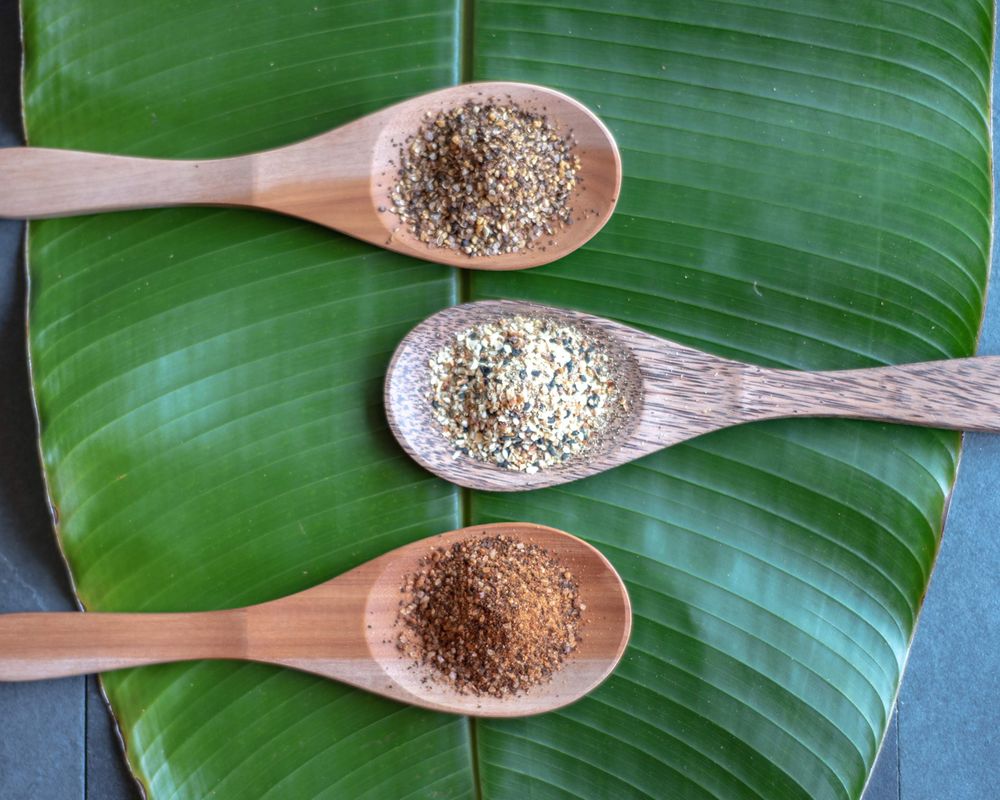 Three wooden spoons with different seasonings resting on a large green banana leaf.